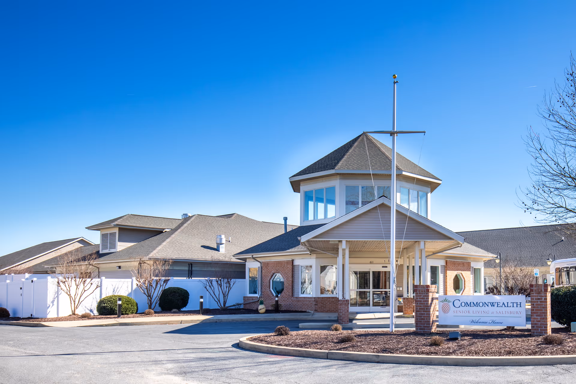 Front entrance of Commonwealth Senior Living at Salisbury showing a brick and siding building with a covered porte-cochere and flagpole under a clear blue sky.