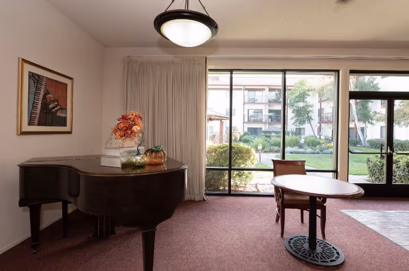 Interior room with a grand piano on the left side decorated with a floral arrangement and a pumpkin ornament. A framed picture of a piano hangs on the wall above the piano. To the right, there is a round table with a single chair. Large floor-to-ceiling windows and a glass door provide a view of a green outdoor garden area with bushes and trees.