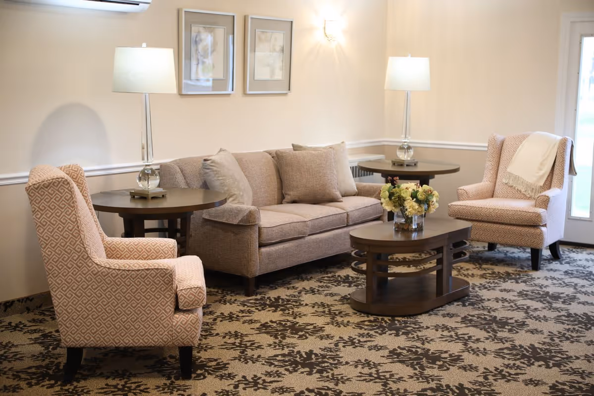 A cozy seating area with a sofa, two upholstered armchairs, coffee and side tables, lamps, and floral centerpiece in a lounge.