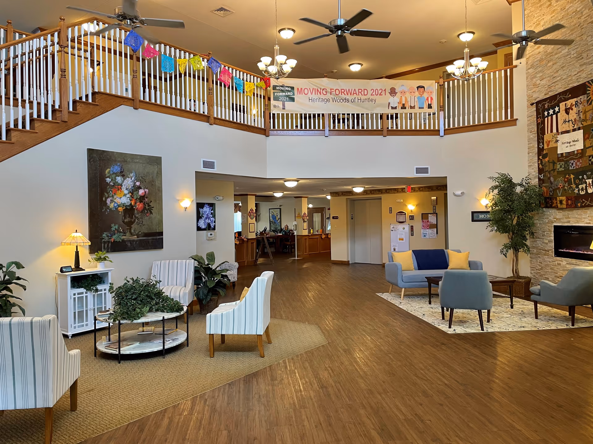 Spacious senior living facility common area with wooden flooring, multiple seating arrangements including striped armchairs and blue sofas with yellow pillows, a stone fireplace on the right wall, ceiling fans, and a second-floor balcony decorated with colorful paper banners and a 'Moving Forward 2021 Heritage Woods of Huntley' banner.