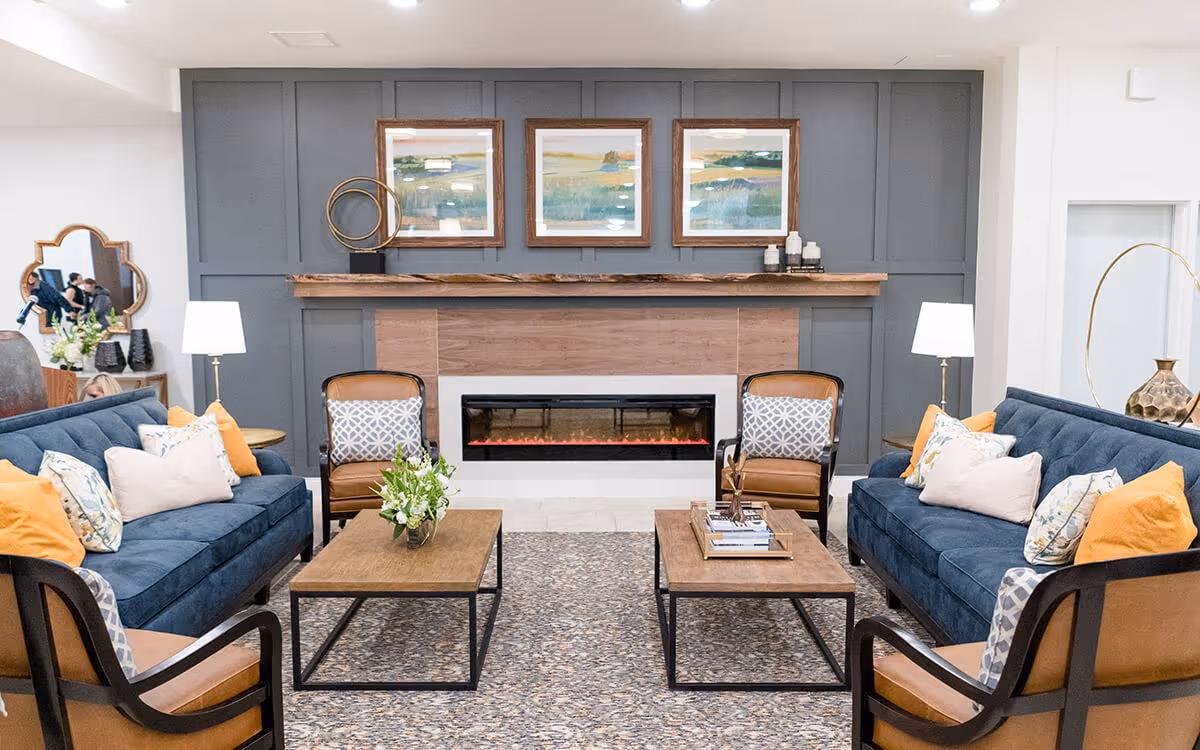 A cozy living room with two blue sofas adorned with yellow and patterned pillows facing each other across two wooden coffee tables. There are two brown armchairs with patterned cushions on either side of the tables. The room features a modern electric fireplace set into a wooden and gray paneled wall, above which hang three framed landscape paintings. Two floor lamps with white shades flank the fireplace, and decorative items including a vase with flowers and sculptural pieces are placed on the tables and mantel.