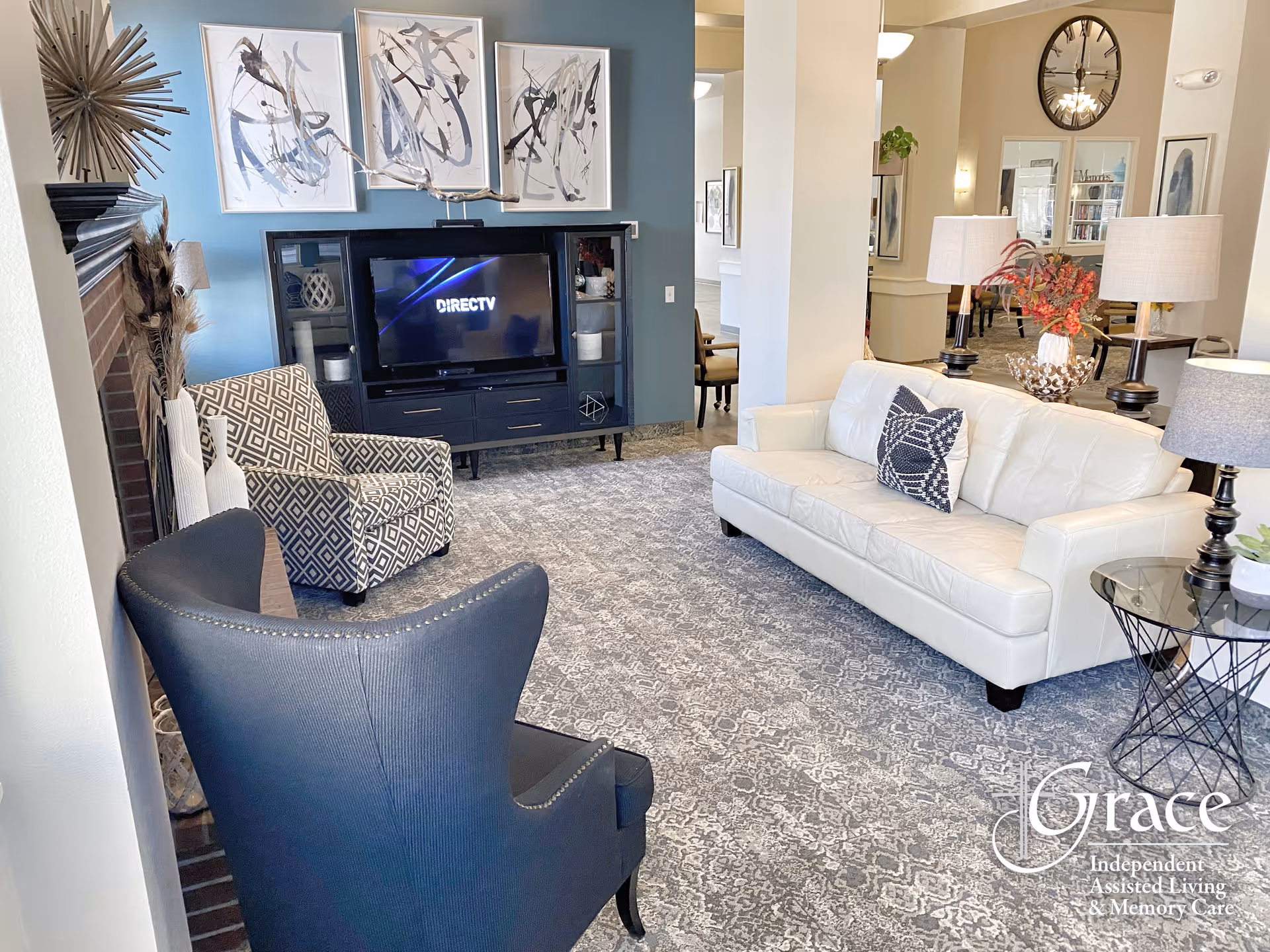 A cozy living room area with a white leather sofa, two armchairs (one patterned and one dark blue), a patterned carpet, a TV on a black entertainment unit, and three abstract paintings on a blue accent wall. There are also two table lamps on side tables and decorative items including a vase with flowers and a wall clock in the background.