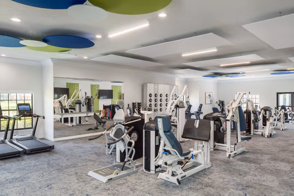A spacious fitness room in Regency Oaks Health Center featuring various exercise machines including treadmills, weight machines, and stationary bikes. The room has large windows allowing natural light, mirrors on the walls, and colorful circular ceiling panels in blue and green.