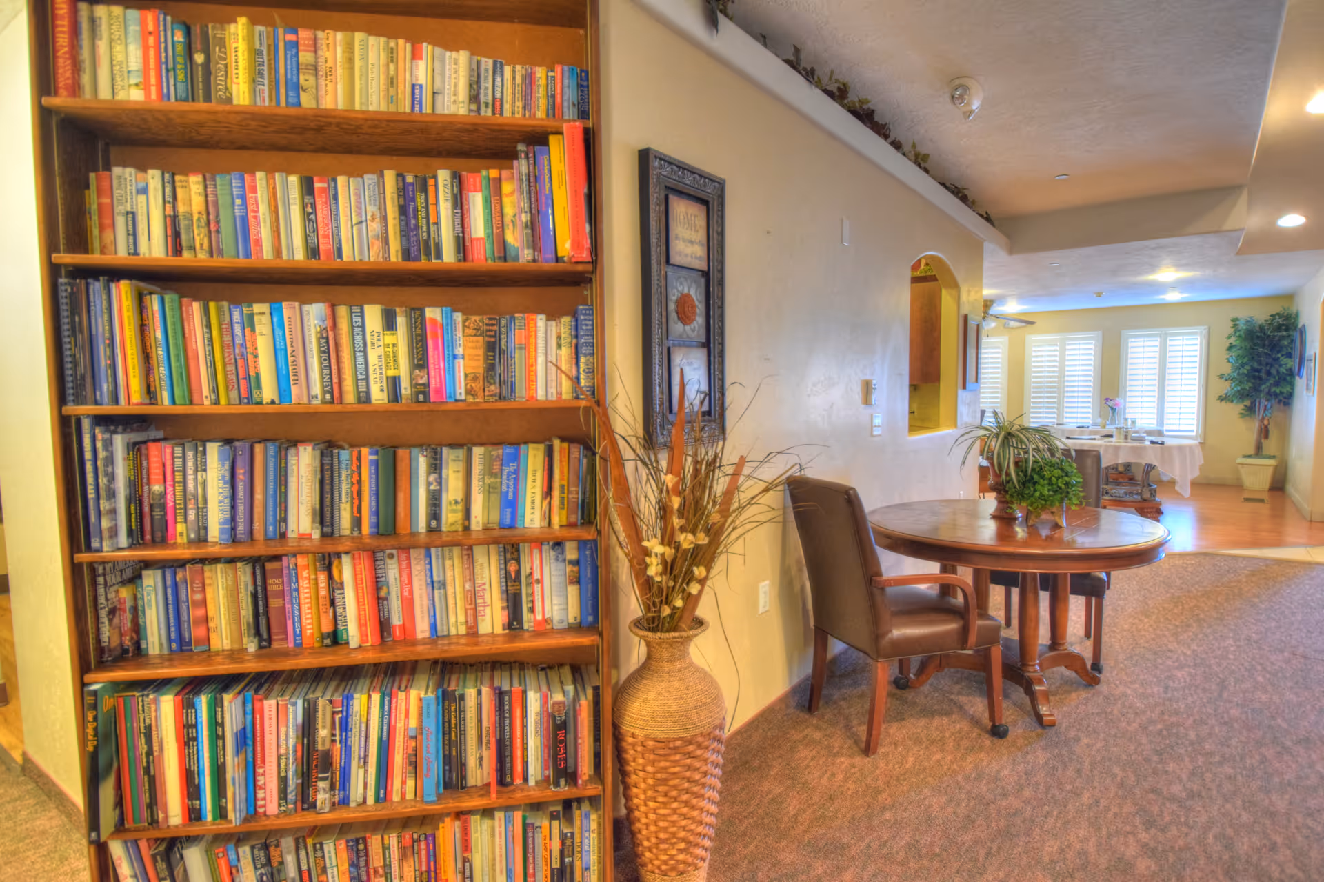 Interior view of a senior living facility showing a tall wooden bookshelf filled with books on the left, a round wooden table with a plant centerpiece and a brown chair next to it, a decorative vase with dried plants, and a hallway leading to a dining area with large windows covered by white shutters and a potted plant in the corner.