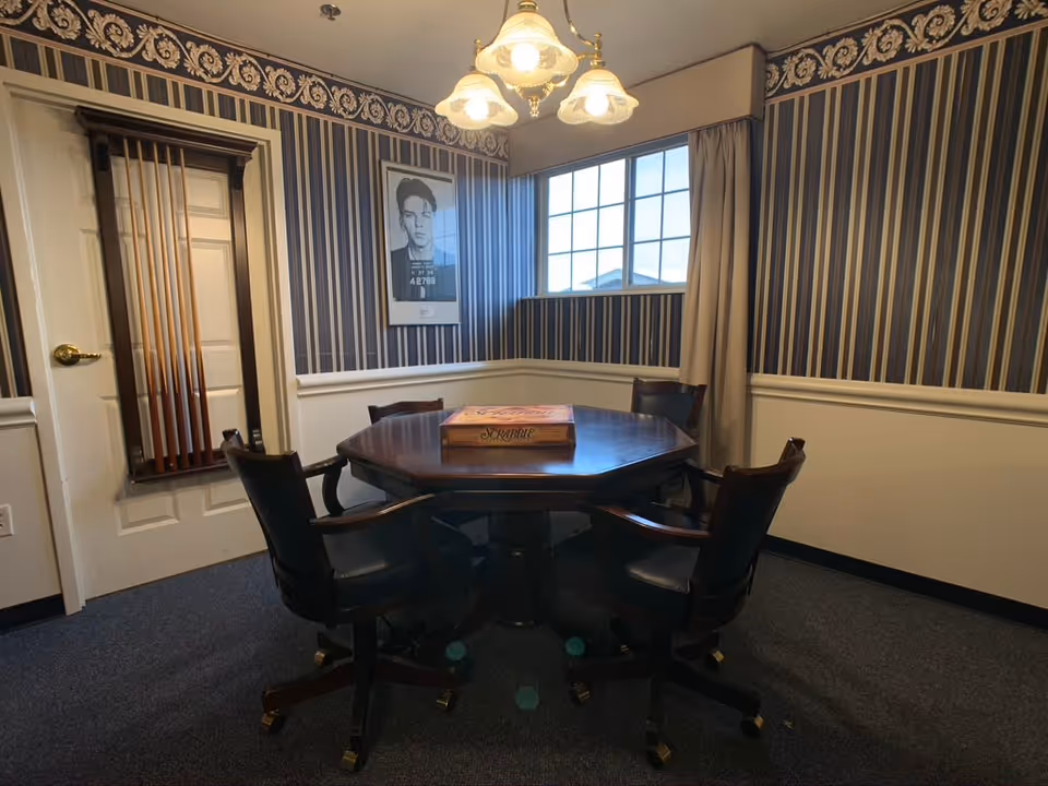 A small room with striped wallpaper and a window with beige curtains. In the center is a dark wooden octagonal table with a Scrabble board game on top, surrounded by four black leather office chairs with wooden armrests and wheels. A wall-mounted rack holding pool cues is visible next to a closed white door. A framed black and white photo hangs on the wall above the table. A ceiling light fixture with three lamps illuminates the room.