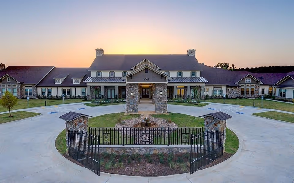 Front exterior view of Ridgemere Senior Living facility at sunset, featuring a large building with stone pillars, a gated entrance, and a circular driveway surrounded by landscaped greenery.