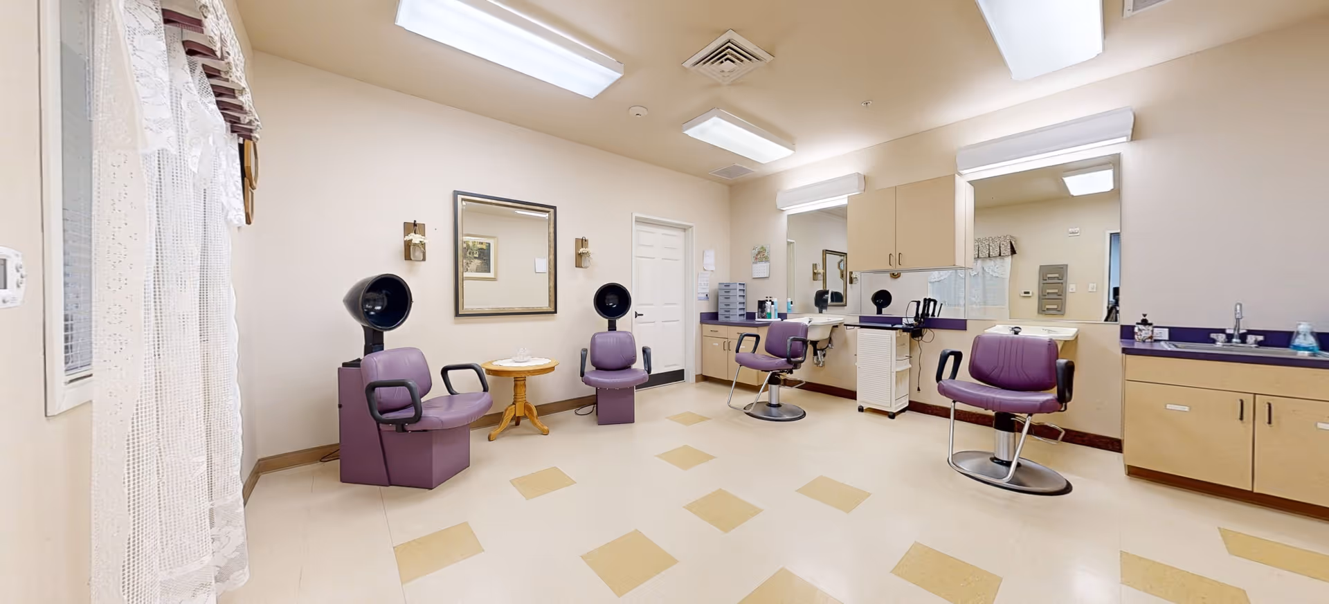 Interior view of a salon area in a senior living facility with three purple salon chairs, two with hair dryers attached. The room has beige walls, a large mirror, cabinets, and a small round table between two chairs. The floor is tiled with a pattern of beige and light brown squares.