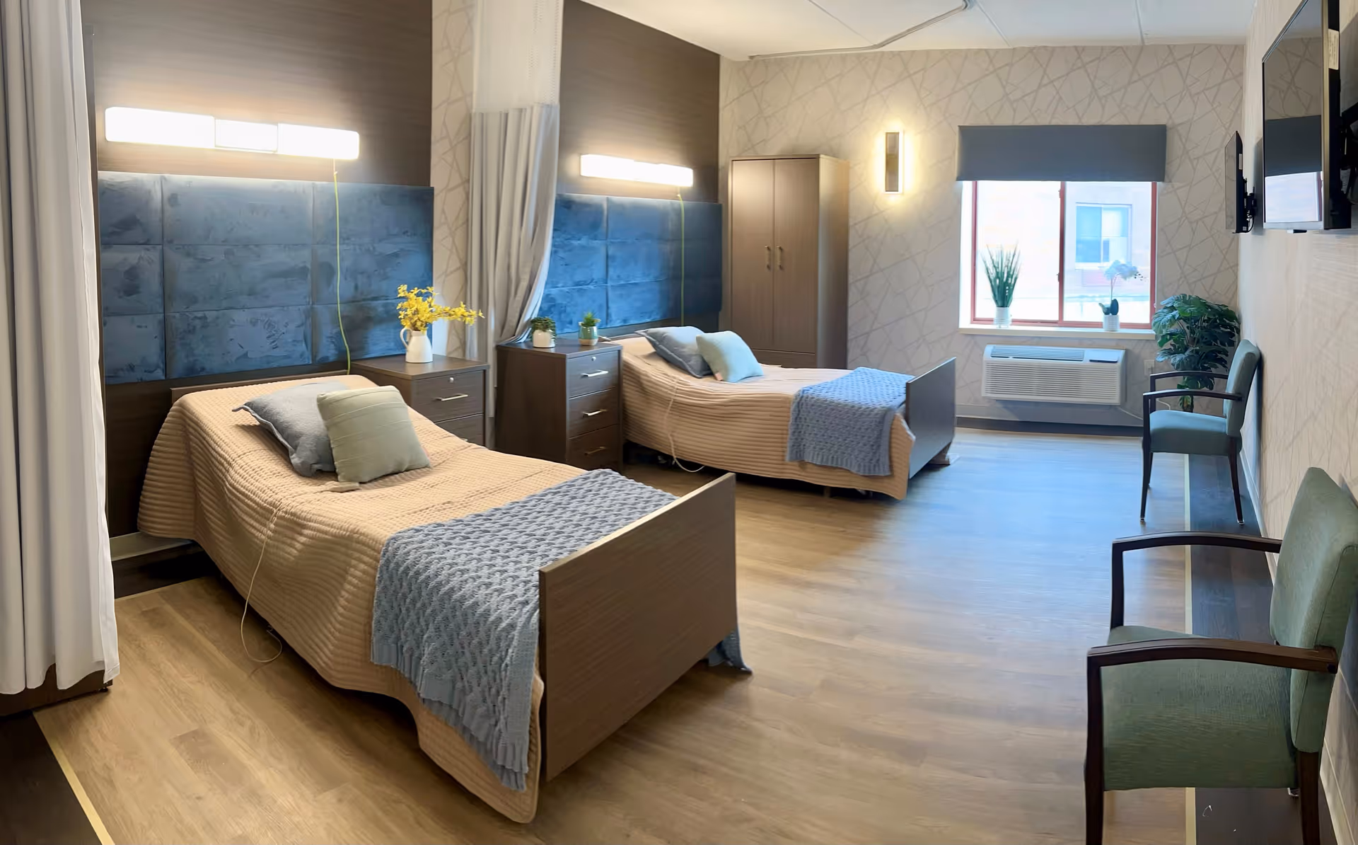 A bright and clean bedroom in a senior living facility with two single beds, each with a beige bedspread and a blue throw blanket. The beds have padded blue headboards and are separated by a small wooden nightstand with a vase of yellow flowers. There are two green chairs along the right wall, a window with a gray shade, and a wall-mounted TV. The room has wooden flooring and soft lighting fixtures on the walls.