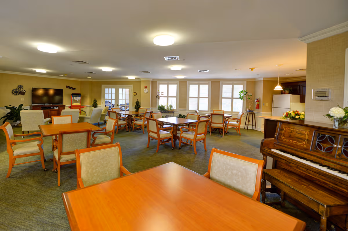 A spacious common area in a senior living facility with multiple wooden tables and cushioned chairs arranged throughout the room. There is a piano on the right side, a television on a stand against the far wall, and a small kitchenette area with a refrigerator and countertop. The room is well-lit with ceiling lights and natural light coming through several windows. Potted plants and flowers add a touch of greenery to the space.