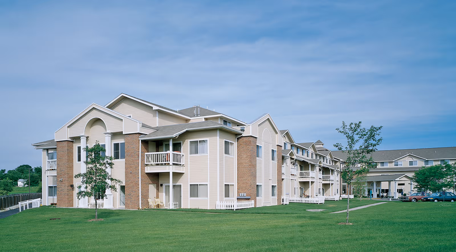 Exterior view of a multi-story senior living building with balconies, brick and siding facade and a large green lawn.