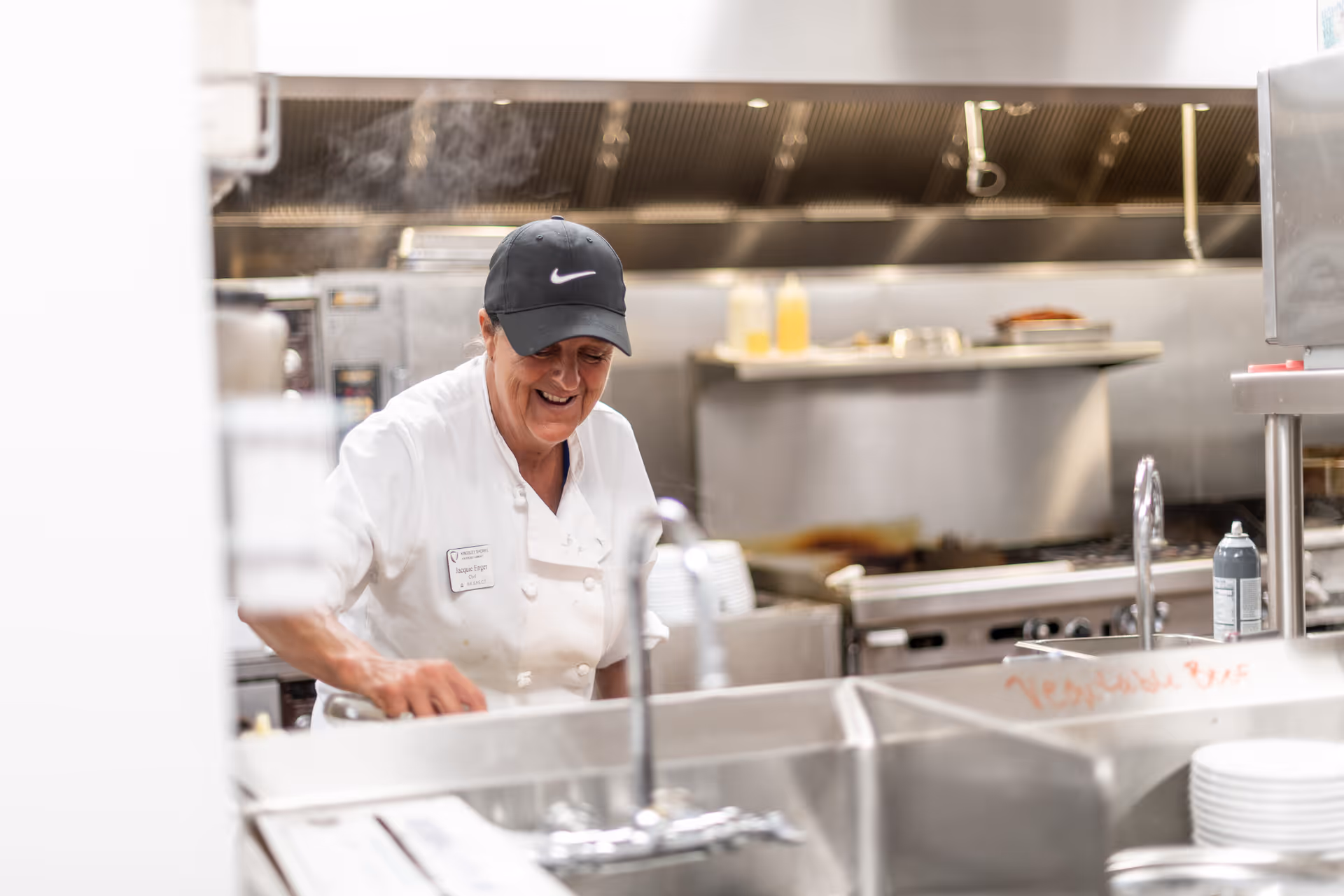 A chef wearing a white uniform and black Nike cap is smiling while working in a commercial kitchen with stainless steel appliances and sinks.