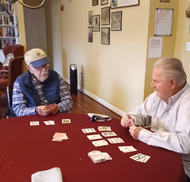 Two elderly men sit at a table playing cards in a communal room with framed photos on the wall.