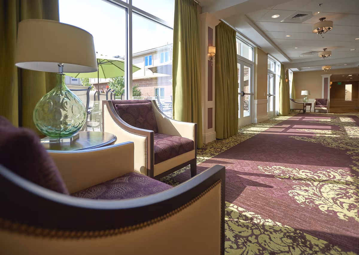 A bright and spacious interior hallway with large windows letting in natural light. The hallway features patterned carpet in purple and green tones, green curtains, and comfortable seating areas with beige and purple upholstered chairs and glass table lamps. Outside the windows, patio umbrellas and part of a building are visible.
