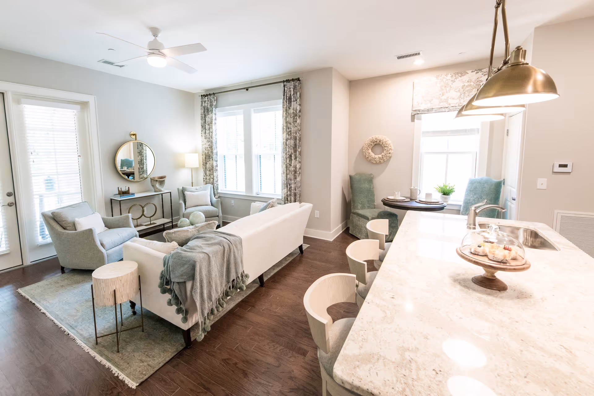Bright and airy living room area with a white sofa draped with a gray throw blanket, two light blue armchairs, a round mirror on the wall above a console table, and large windows with patterned curtains. Adjacent to the living room is a kitchen island with marble countertop, three modern bar stools, pendant lights, and a small round dining table with two green upholstered chairs near a window.