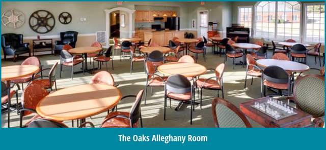 Spacious dining/activity room with multiple round tables and chairs, a kitchen area in back, and large windows.