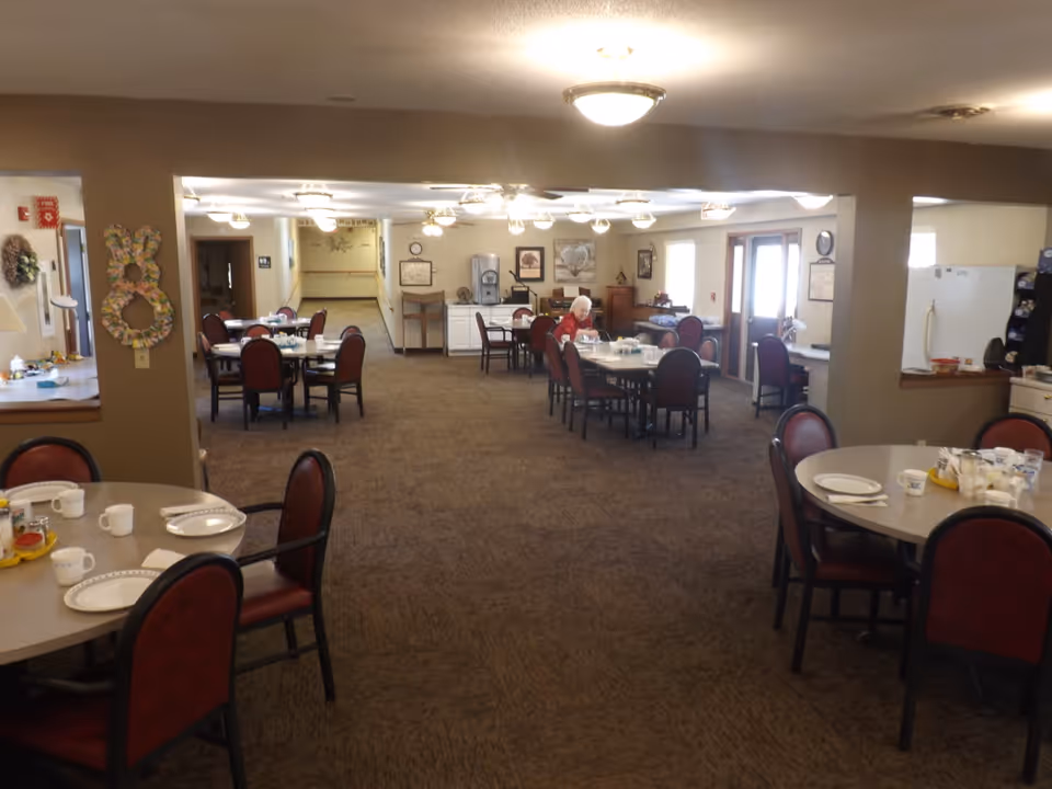 Interior view of a dining room in an assisted living facility with multiple round tables and chairs. One elderly person is seated at a table in the background. The room is well-lit with ceiling lights and has a carpeted floor. There are decorations on the walls and a water dispenser in the back.