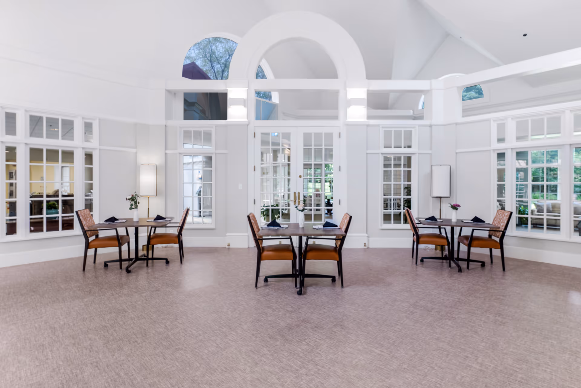 A bright and spacious dining area with three small tables, each set with two chairs and table settings including napkins and small flower arrangements. The room features large windows with white frames, high ceilings with arched windows, and light-colored walls and flooring.