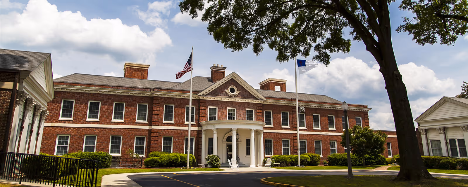 Front exterior view of a large, two-story brick building with white columns at the entrance, two flagpoles with flags, and well-maintained landscaping including bushes and trees under a partly cloudy sky.