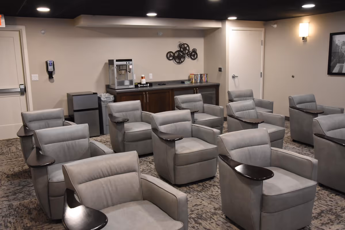 A room with multiple gray upholstered armchairs arranged in rows, each with a small attached wooden side table. At the back of the room, there is a counter with a popcorn machine, a small refrigerator, and some DVDs. The walls are beige with minimal decoration, including a wall-mounted light and a circular metal wall art piece.