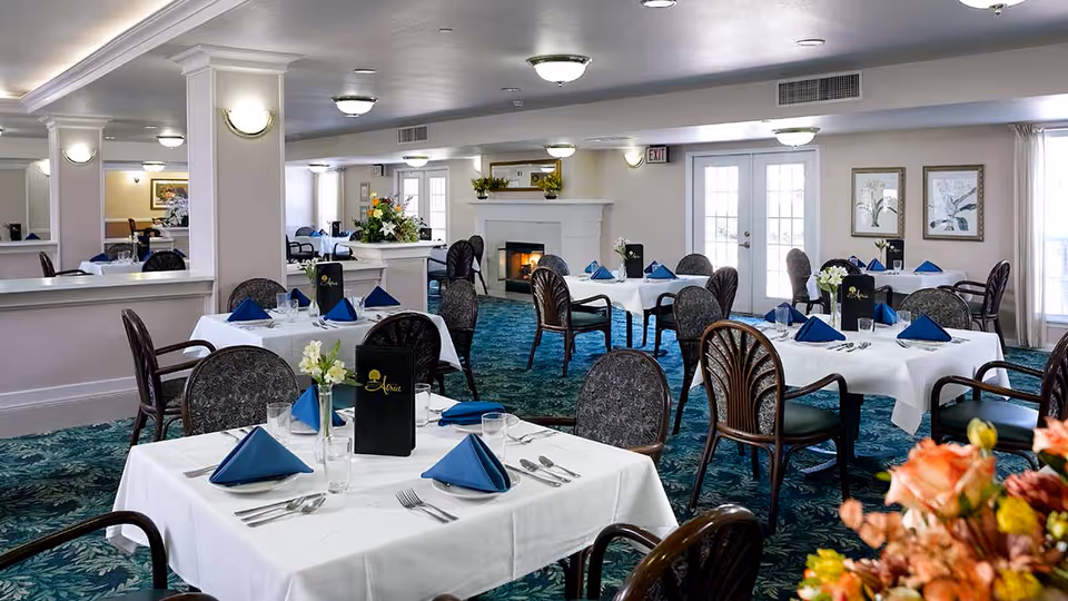 A dining room in a senior living facility with multiple tables covered in white tablecloths, each set with blue folded napkins, silverware, glasses, and small flower arrangements. The room has patterned carpet, several chairs around each table, a fireplace in the background, and soft lighting from ceiling fixtures and wall sconces.