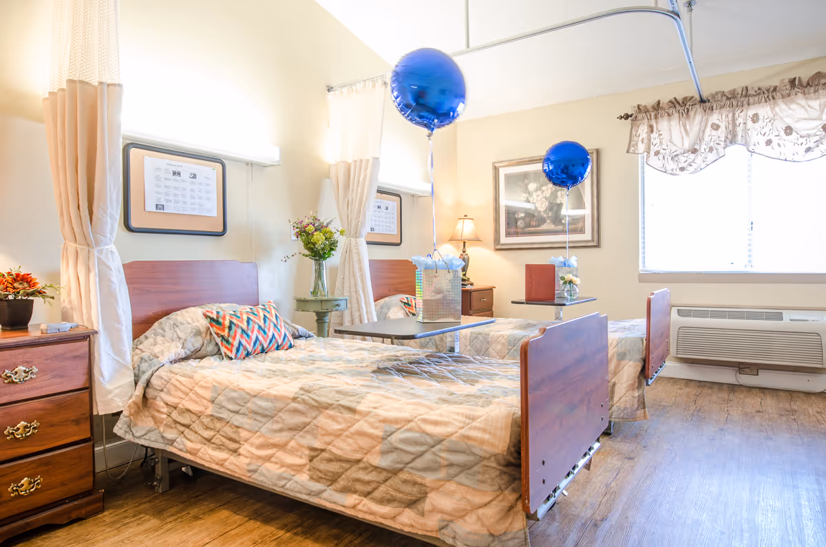 A bright and clean shared bedroom in a senior living facility with two single beds, each with a quilted bedspread and a decorative pillow. There are blue balloons tied to a small table positioned between the beds. The room has wooden furniture including nightstands and a small table with flowers and a lamp. A large window with floral curtains lets in natural light, and the floor is wooden.