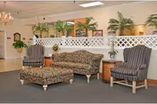 A seating area in a senior living facility featuring a floral patterned sofa with a matching ottoman, two striped armchairs, and a wooden side table. The background includes a white lattice partition with potted plants on top and a chandelier hanging from the ceiling.