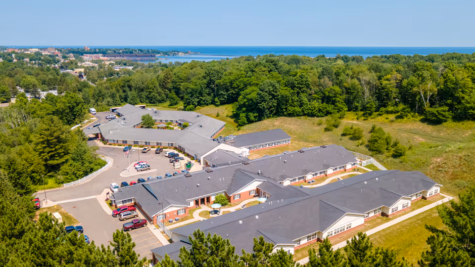 Aerial view of Brookridge Heights senior living facility surrounded by trees and greenery, with a parking lot and multiple connected buildings under a clear blue sky and a body of water visible in the background.