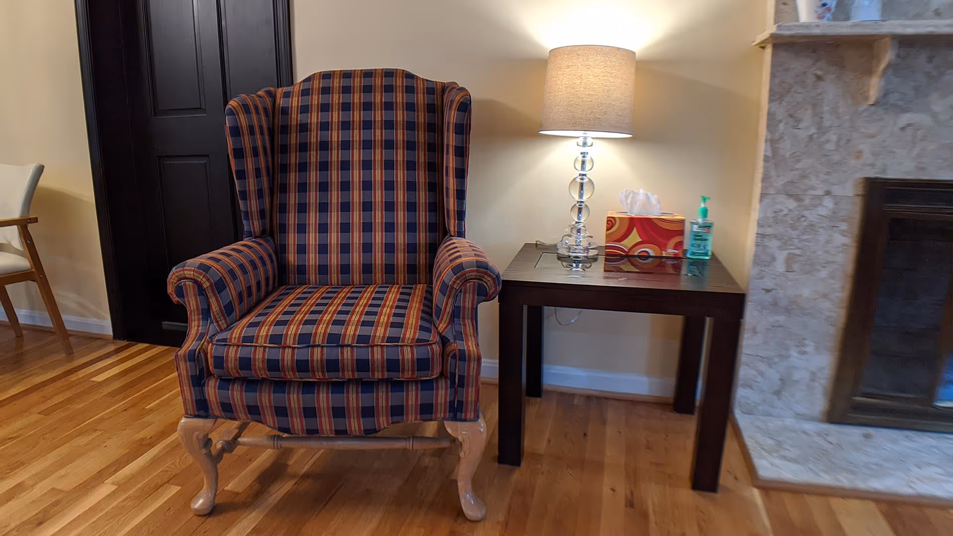 A cozy living room corner featuring a plaid upholstered armchair with wooden legs, a dark wooden side table with a glass lamp, a box of tissues, and a bottle of hand sanitizer. To the right is a marble fireplace, and to the left, part of another chair and a dark door are visible. The floor is wooden.