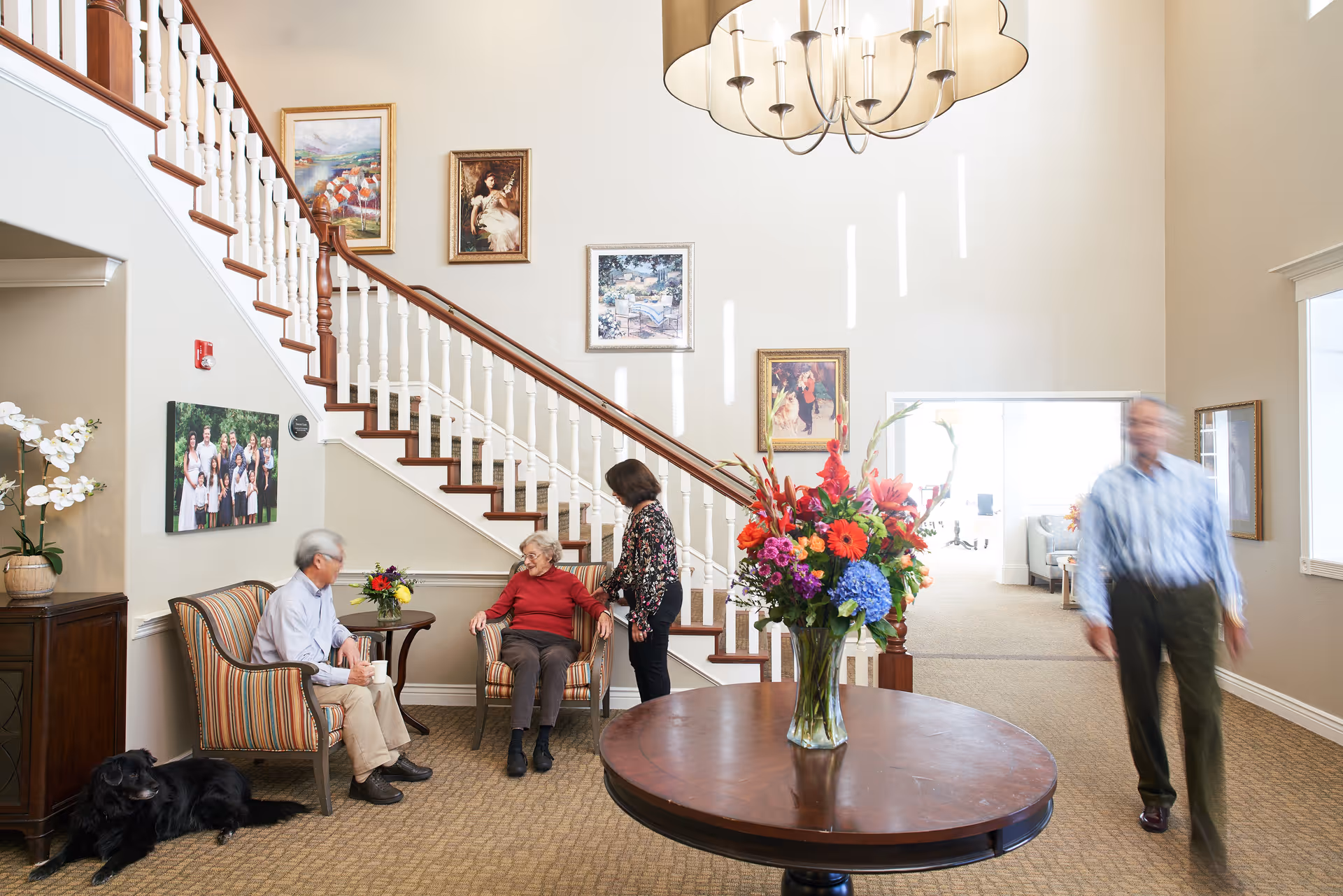A bright and spacious living area in a senior living facility with a staircase and several framed paintings on the wall. Three elderly people are seated and standing near the staircase, engaged in conversation, while a man walks by. A large round wooden table with a colorful flower arrangement is in the foreground, and a black dog lies on the carpet near a wooden cabinet with a potted orchid.