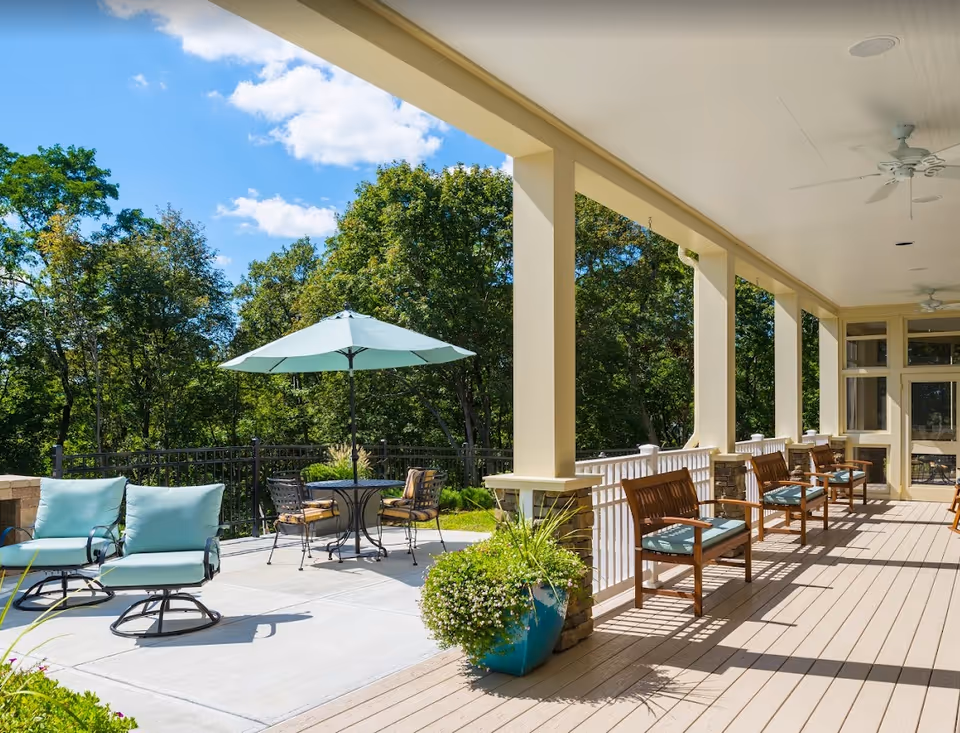 A spacious outdoor patio area at Brightview Arlington Senior Assisted Living and Memory Care, featuring several wooden chairs with blue cushions lined up along a covered porch with ceiling fans. In the open area, there are cushioned lounge chairs and a round table with an umbrella surrounded by metal chairs. The patio overlooks a green landscape with trees under a blue sky with some clouds.