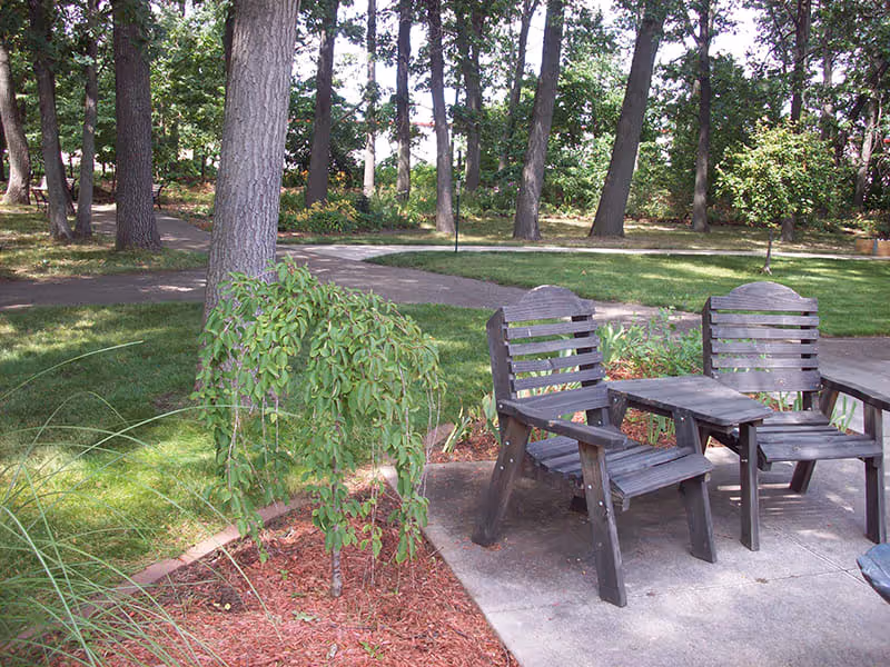 Outdoor patio area with two wooden chairs and a small wooden table on a concrete surface, surrounded by green grass, trees, and landscaped plants in a garden setting.