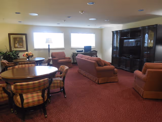 A senior living community common room with red carpet and beige walls. The room contains a round wooden table with four plaid upholstered chairs, two armchairs, a sofa, a floor lamp, a desk with a computer, and a large dark wooden entertainment center with a TV. Two windows provide natural light.