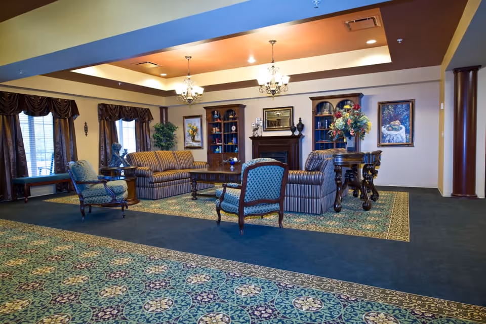 A spacious and elegantly decorated living room with patterned carpet, two striped sofas, two blue upholstered armchairs, a wooden coffee table, and a side table with a large floral arrangement. The room features a fireplace with a mirror above it, two wooden bookshelves, framed paintings on the walls, and large windows with dark curtains. The ceiling has recessed lighting and two chandeliers.