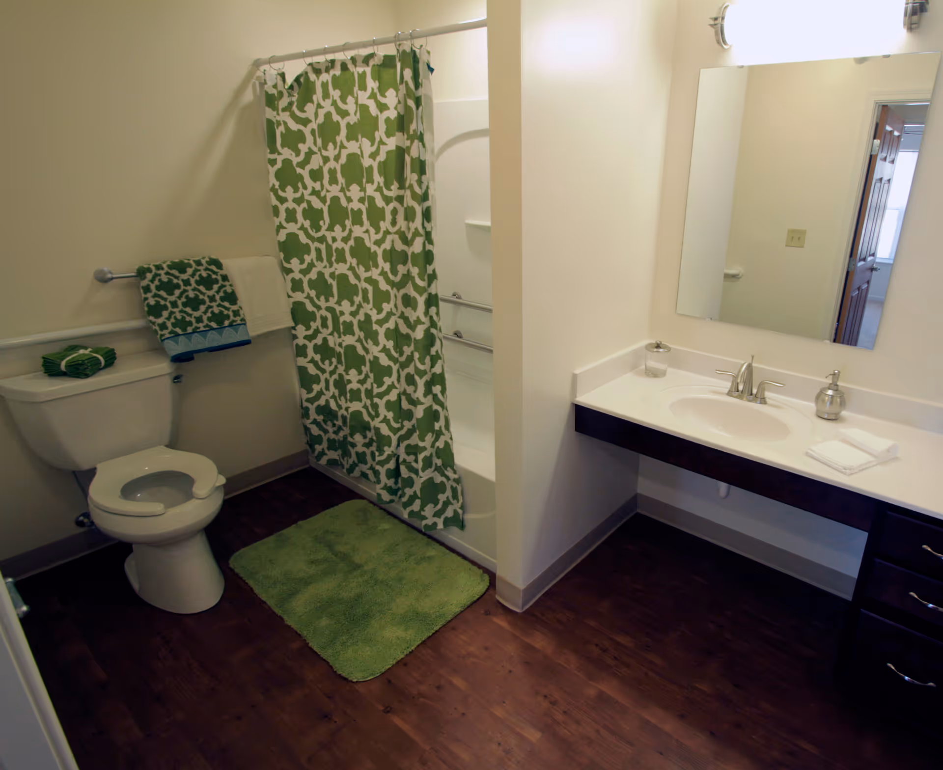 Bathroom with a toilet, a green and white patterned shower curtain, a green bath mat, and a sink with a large mirror above it. Towels are neatly placed on the toilet tank and hanging on a towel rack.