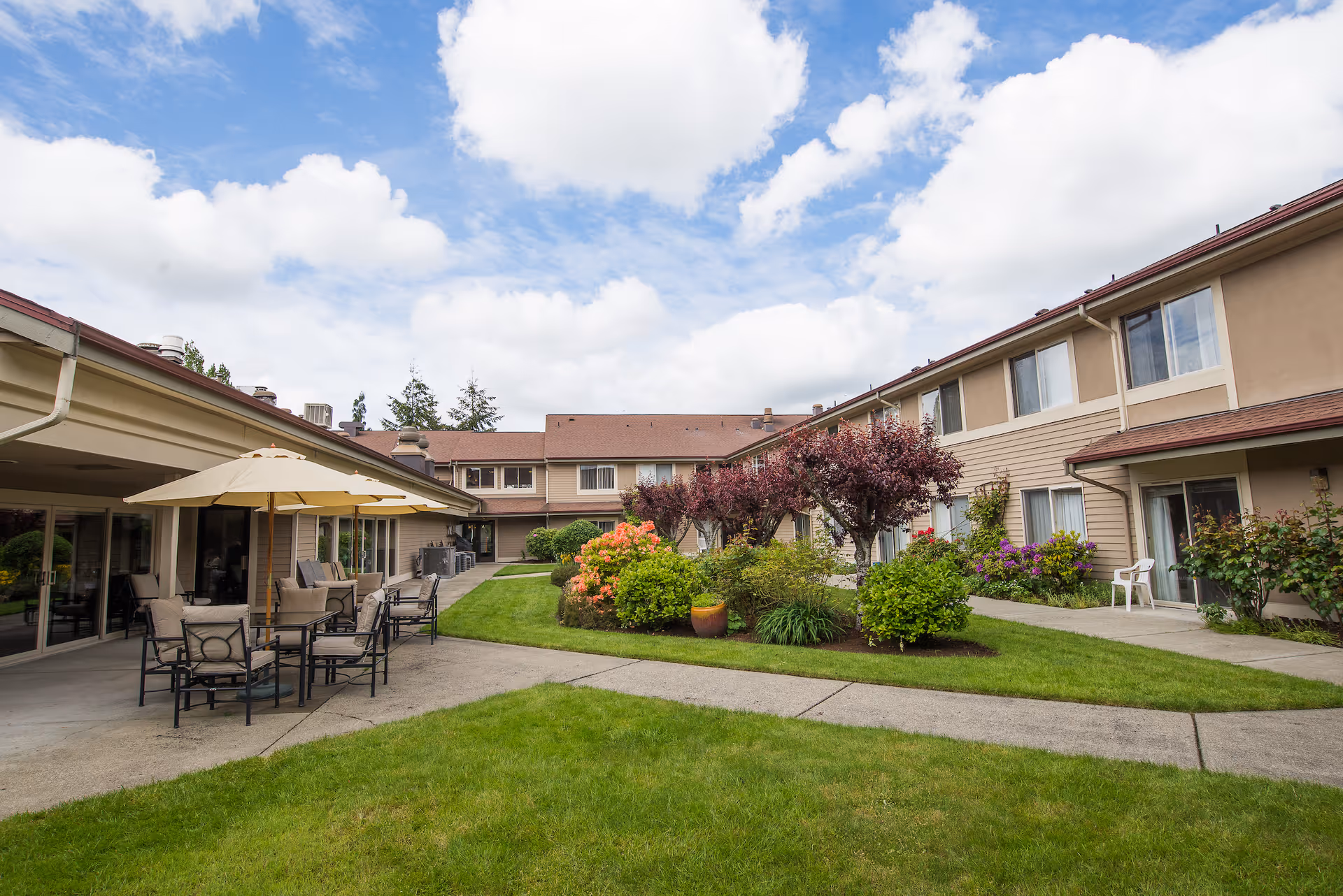 Outdoor courtyard area of a senior living facility with green grass, landscaped bushes, and small trees. There are patio tables with umbrellas and chairs on a concrete patio next to the building. The building is two stories with beige siding and multiple windows. The sky is partly cloudy.