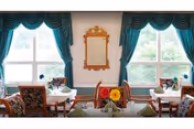Bright dining room with multiple set tables, floral centerpieces, teal window drapes, and a decorative mirror on the wall.