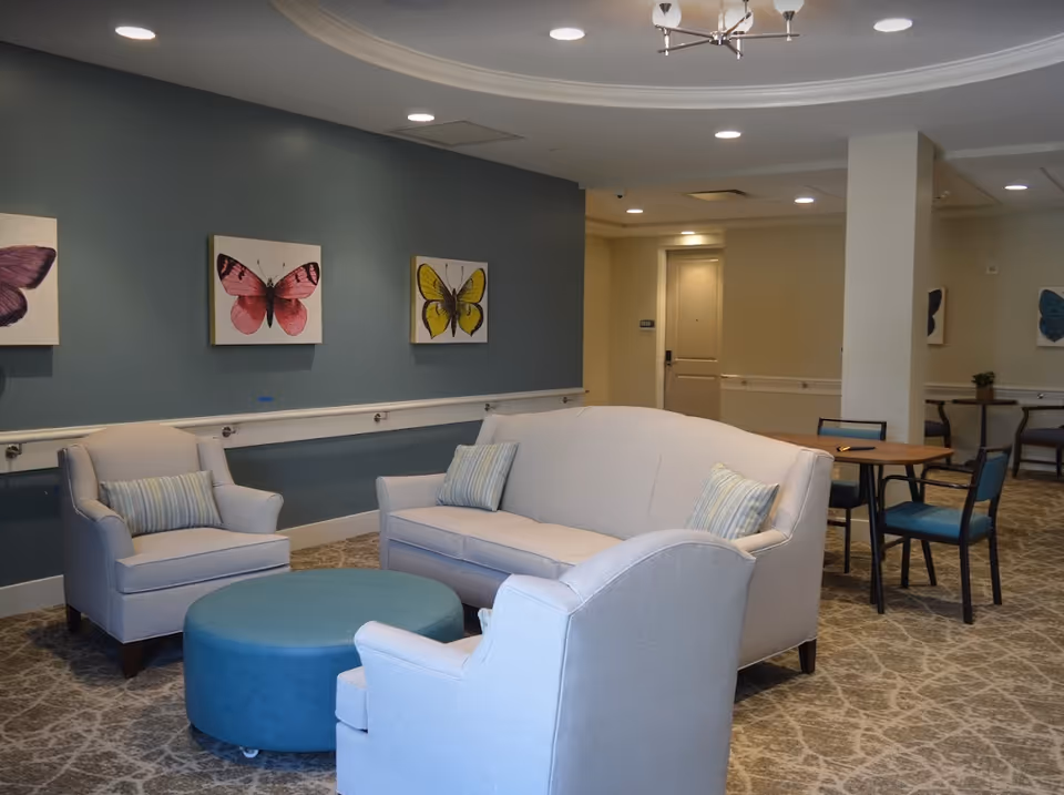 A cozy sitting area in a senior living facility with a light gray sofa and two matching armchairs arranged around a round blue ottoman. The walls are painted blue and decorated with framed butterfly artwork. In the background, there are tables and chairs, beige walls, and a door with the number 1030.