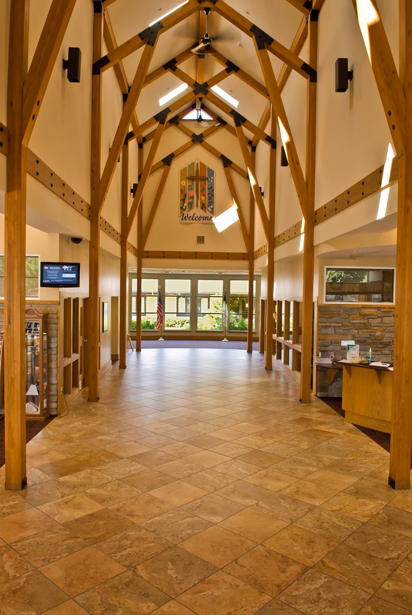 Spacious interior hallway with high vaulted ceiling supported by wooden beams, tiled floor, and large windows at the far end letting in natural light. A welcome sign with a cross and floral design hangs on the far wall. There is a small reception desk on the right side and an American flag near the windows.