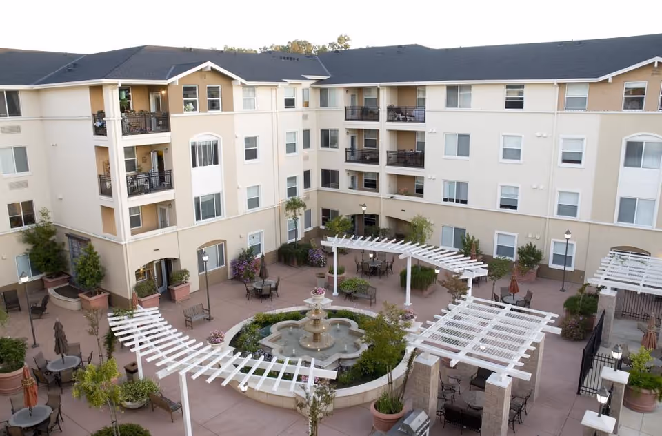 Outdoor courtyard area of a senior living facility with a central water fountain surrounded by seating areas, tables with umbrellas, potted plants, and white pergolas. The courtyard is enclosed by a multi-story beige building with balconies and windows.