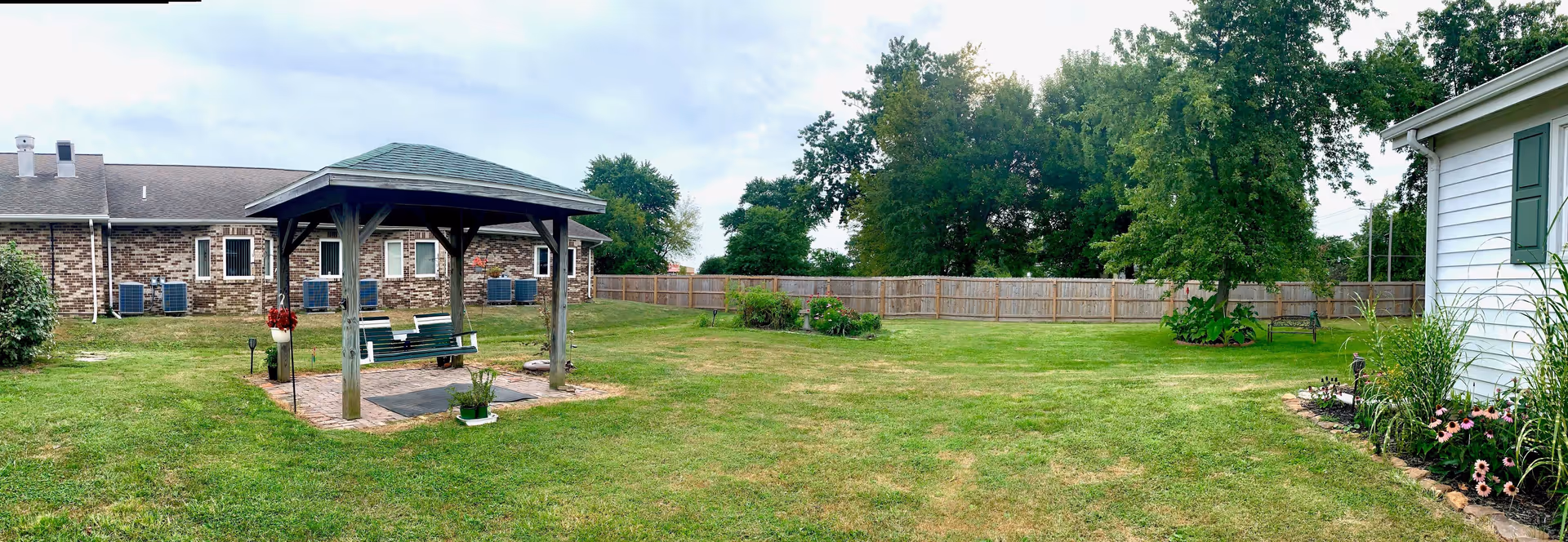 A spacious outdoor garden area at The Oaks Manor featuring a wooden gazebo with a green bench swing underneath. The garden is surrounded by a wooden fence and has well-maintained grass, flower beds, and several trees. A brick building with multiple windows and air conditioning units is visible on the left side, and a white building with green shutters is on the right.