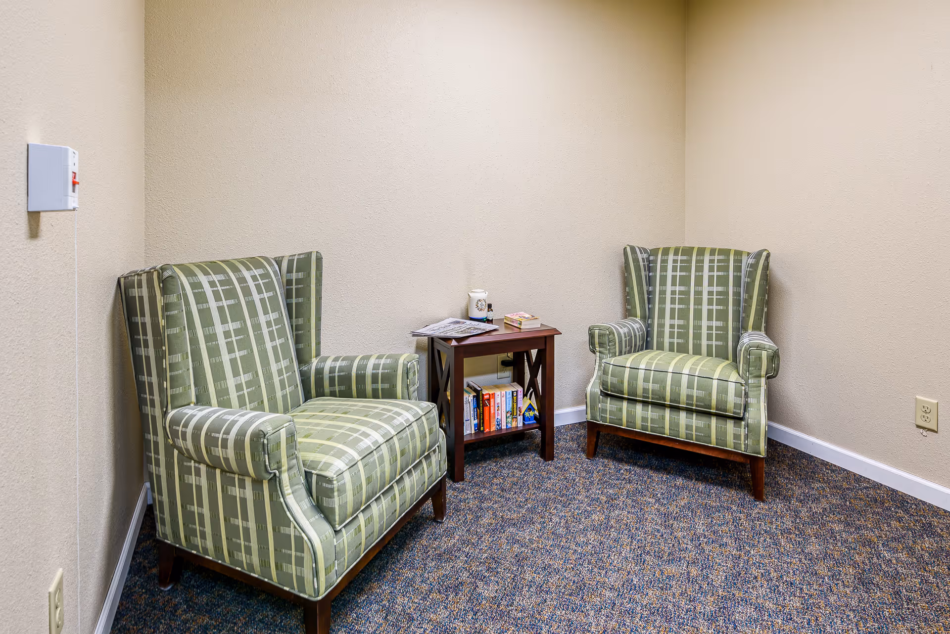 A small seating area with two green plaid upholstered armchairs facing each other, separated by a wooden side table. The table holds a few books, a newspaper, a small decorative jar, and a small bottle. The room has beige walls and a carpeted floor with a blue and brown pattern.