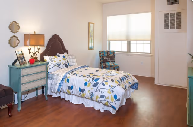 A bright bedroom with a single bed featuring floral and leaf patterned bedding. Next to the bed is a teal nightstand with a lamp, a framed photo, and a red apple decoration. There is a patterned armchair near a window with a white blind, and a framed picture on the wall. The floor is wooden, and the walls are painted white.