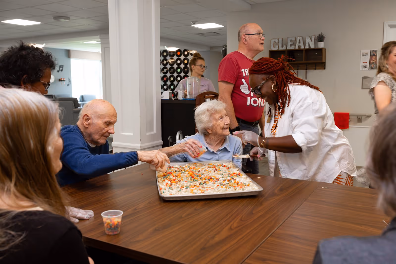A group of elderly people and a caregiver gathered around a table in a communal room, engaging in a cooking or baking activity with a large tray of food. The caregiver is smiling and interacting with an elderly woman, while others participate or watch. The room has a cozy atmosphere with decorations and furniture in the background.