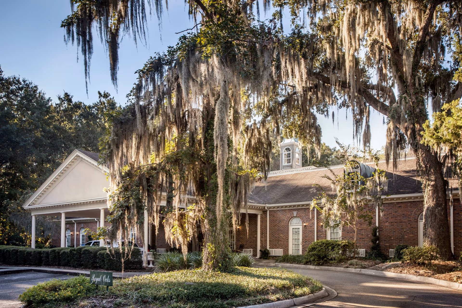 Brick senior living building with a columned entrance, circular driveway, and large moss-draped oak trees.
