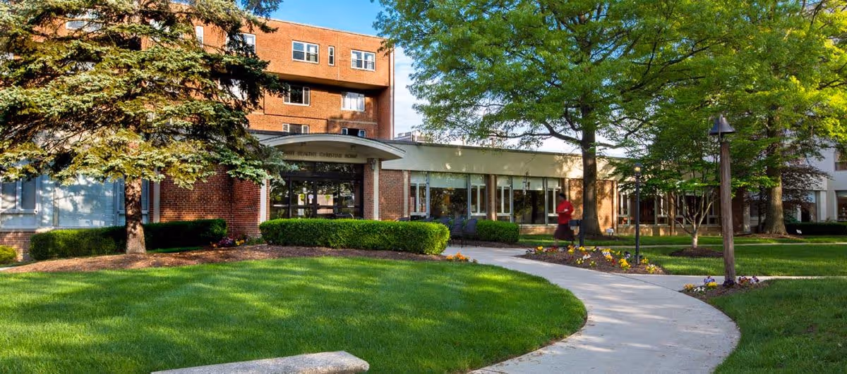 Exterior view of The Christian Village at Mt Healthy building with a curved concrete walkway, green lawn, trees, shrubs, and flower beds in the foreground. A person is walking near the entrance of the building.