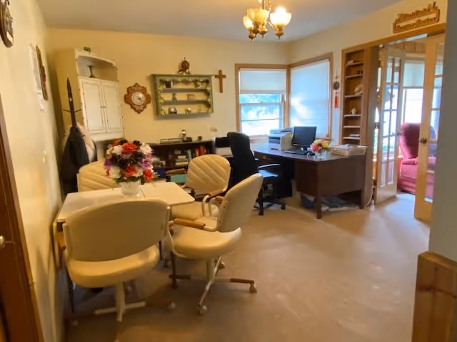 A cozy office and sitting area in a senior living facility with a desk, computer, office chair, and a small table with four cushioned chairs. There are flowers on the table, a bookshelf with decorative items, a wall clock, and a cross on the wall. A doorway with glass panels leads to another room with a red armchair.