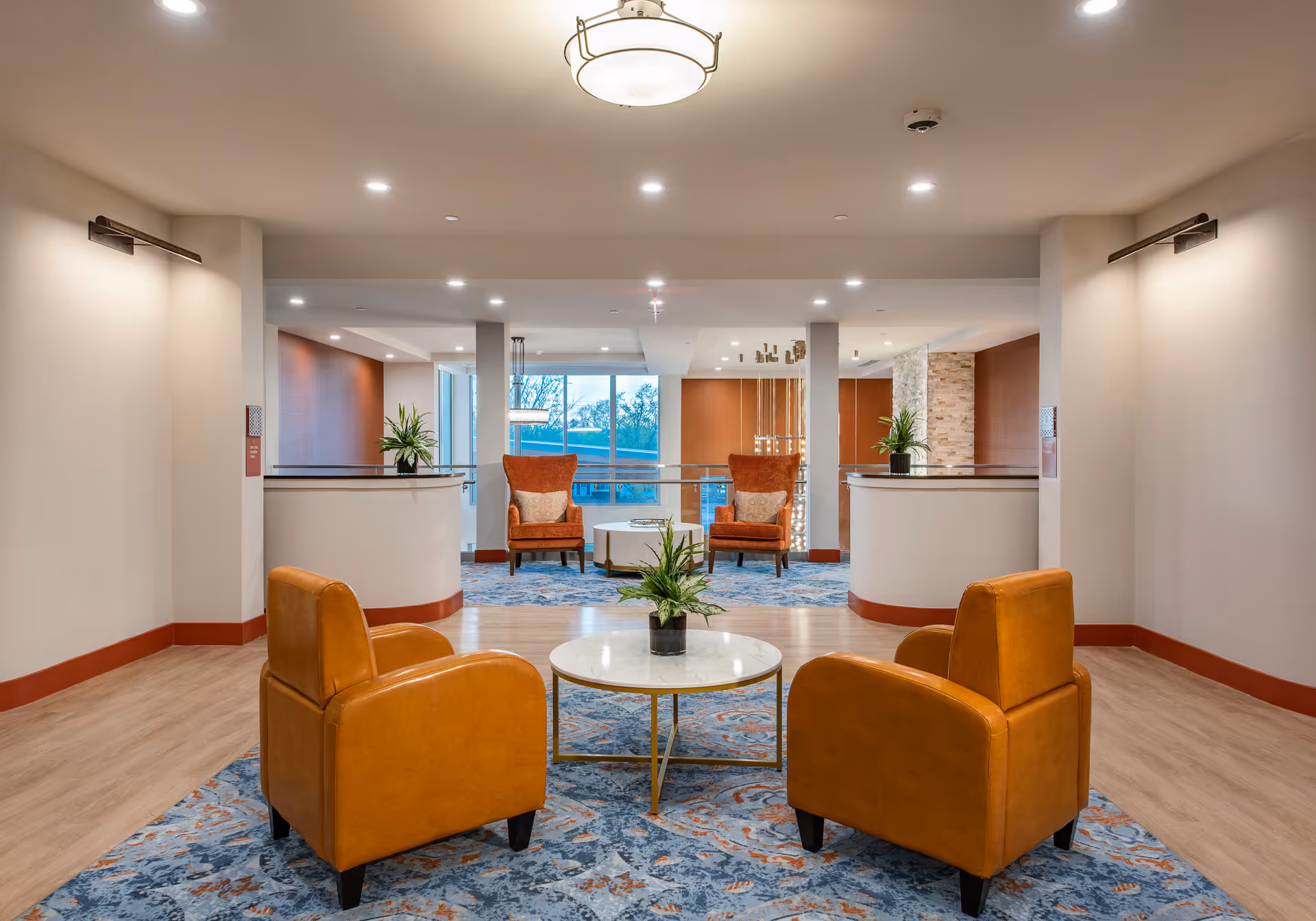 Bright, modern lounge area with orange leather armchairs arranged around a round coffee table, plants, and reception desks in the background.
