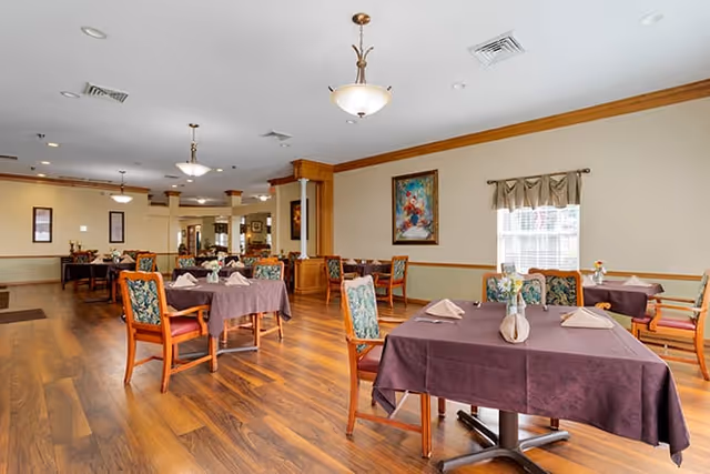 A spacious dining room with several tables covered in purple tablecloths, each set with folded napkins and small floral centerpieces. The room has wooden flooring, beige walls with a wooden trim, framed artwork, and windows with curtains allowing natural light to enter. Comfortable chairs with floral upholstery surround the tables.