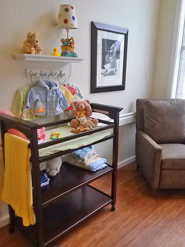 A cozy corner of a room featuring a dark wooden changing table with folded blankets and stuffed animals on its shelves. Above the table, a small white shelf holds a teddy bear, a rubber duck, and a lamp with a colorful polka dot shade. Several baby clothes hang on hooks beneath the shelf. To the right, there is a gray upholstered armchair and a framed black and white photo of a baby on the wall. The room has wooden flooring and a window letting in natural light.