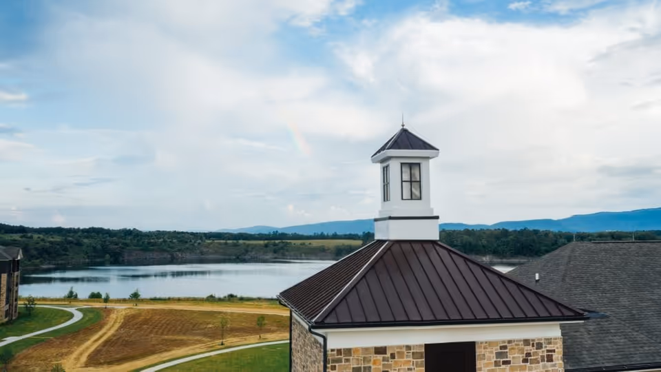 View of a senior living facility building with a stone exterior and a dark metal roof topped with a small white cupola. In the background, there is a large body of water, green fields, trees, and distant mountains under a partly cloudy sky with a faint rainbow.